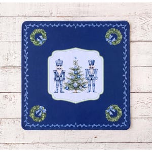 May include: A square navy blue fabric mat featuring a central white scalloped frame containing a green Christmas tree flanked by two blue nutcrackers. The corners display green wreaths with blue bows, and a vine border frames the entire edge.