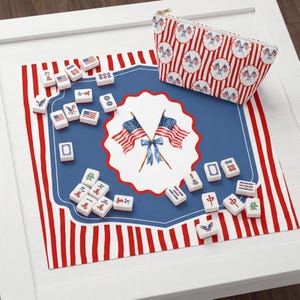 May include: A Mahjong set with a patriotic theme, featuring red, white, and blue colours. The set includes tiles, a patterned mat, and a matching zipped pouch. The mat displays American flags and a scalloped blue border.