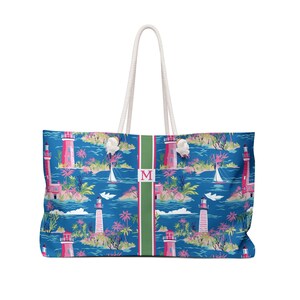May include: A tote bag featuring a blue background with a repeating pattern of pink lighthouses, palm trees, and sailboats. A vertical green stripe runs down the center with a pink letter M inside a white square. It has two white rope handles.
