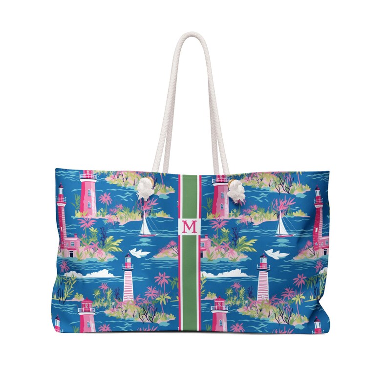 May include: A tote bag featuring a blue background with a repeating pattern of pink lighthouses, palm trees, and sailboats. A vertical green stripe runs down the center with a pink letter M inside a white square. It has two white rope handles.