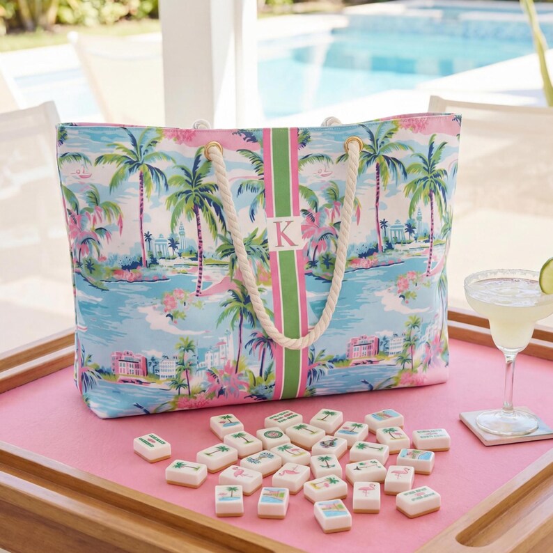 May include: A rectangular tote bag featuring a tropical palm tree and coastal building print in shades of blue, pink, and green. The bag has a central vertical stripe with a pink letter K and thick white rope handles with gold-tone metal hardware.