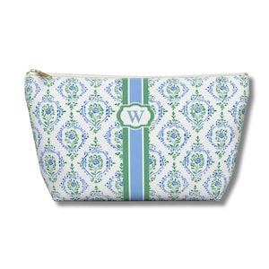 May include: A white cosmetic bag with a blue and green floral pattern. A green and blue stripe runs down the center, with a white oval containing the letter "W". The bag has a gold zipper.