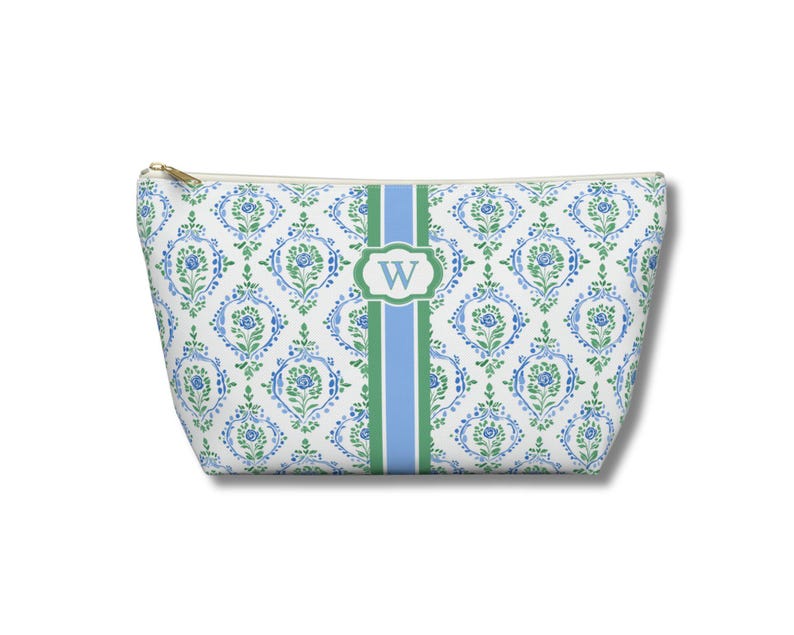 May include: A white cosmetic bag with a blue and green floral pattern. A green and blue stripe runs down the center, with a white oval containing the letter "W". The bag has a gold zipper.