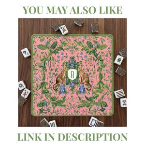 May include: Pink mahjong mat with a green border, featuring a botanical design with two cheetahs, blue vases, and the letter "R". The mat is surrounded by mahjong tiles. The text "YOU MAY ALSO LIKE" and "LINK IN DESCRIPTION" are also visible.