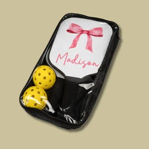 May include: A black zippered carrying case with a clear plastic window showing a white paddleball racket with a pink watercolor bow and the name "Madison" written in pink. Two yellow paddleballs are also visible in the case.