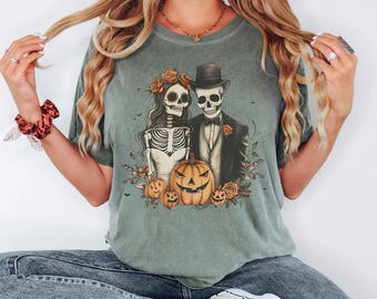May include: A sage green t-shirt featuring a graphic print of two skeletons dressed as a bride and groom, wearing a top hat and floral crown, surrounded by orange pumpkins and autumn flowers. The design is centered on the chest of the garment.