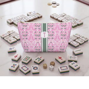 May include: A pink zippered pouch featuring a white and green floral damask pattern with a central vertical green stripe and the letter T. The pouch is surrounded by numerous rectangular mahjong tiles and three small dice on a white surface.