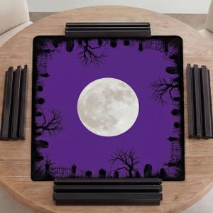 K&ouml;nnte beinhalten: Eine quadratische Spielmatte mit einem leuchtend violetten Hintergrund und einem gro&szlig;en, zentrierten wei&szlig;en Vollmond. Der Rand ist mit einem schwarzen Silhouettenmuster eines Friedhofs verziert, inklusive Grabsteinen, kahlen B&auml;umen und Eisenz&auml;unen.