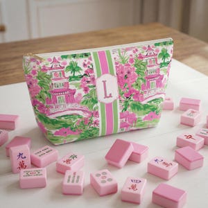 May include: A patterned cosmetic bag with a pink, green, and white floral design, featuring a pink pagoda and the letter "L". Pink Mahjong tiles are scattered around the bag.