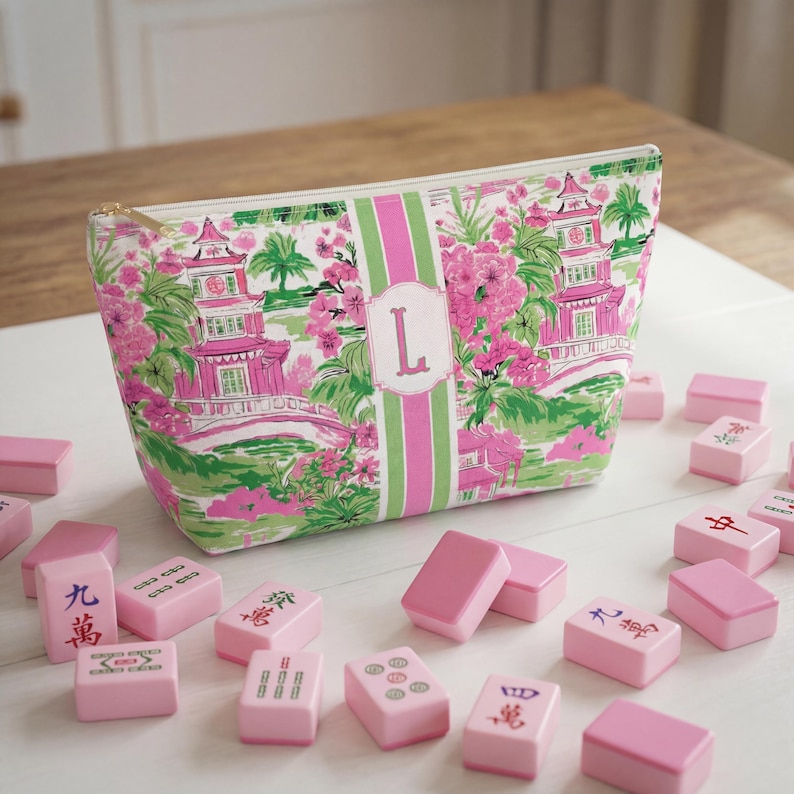 May include: A patterned cosmetic bag with a pink, green, and white floral design, featuring a pink pagoda and the letter "L". Pink Mahjong tiles are scattered around the bag.