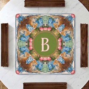 Puede incluir: Mantel individual decorativo cuadrado con un dise&ntilde;o de tem&aacute;tica selv&aacute;tica. La alfombrilla presenta un c&iacute;rculo verde central con la letra "B" en un tono rosa claro. El borde muestra leopardos, patrones florales azules y flores rosas.