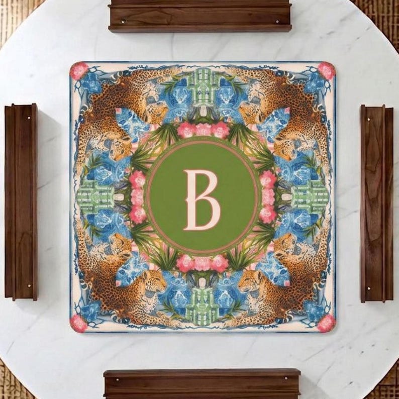 Puede incluir: Mantel individual decorativo cuadrado con un dise&ntilde;o de tem&aacute;tica selv&aacute;tica. La alfombrilla presenta un c&iacute;rculo verde central con la letra "B" en un tono rosa claro. El borde muestra leopardos, patrones florales azules y flores rosas.