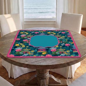 Op de afbeelding: Een vierkante tafelmat met een donkergroenblauwe achtergrond met een herhalend patroon van roze bloemen, gele en blauwe pauwen en rode pagodes. De mat heeft een felroze rand en een groot, effen groenblauw kader in het midden.