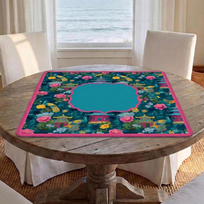 Op de afbeelding: Een vierkante tafelmat met een donkergroenblauwe achtergrond met een herhalend patroon van roze bloemen, gele en blauwe pauwen en rode pagodes. De mat heeft een felroze rand en een groot, effen groenblauw kader in het midden.