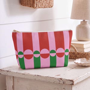 Könnte beinhalten: Eine Reißverschlusstasche mit einem sich wiederholenden geometrischen Muster aus vertikalen rosa und roten Streifen, durchsetzt mit abwechselnden rosa und grünen Kreisen. Die Tasche hat einen goldfarbenen Metallreißverschluss und eine rechteckige Form.