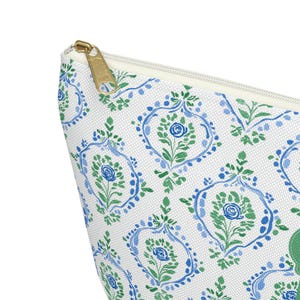 May include: A white cosmetic bag with a repeating pattern of blue and green floral designs. The bag has a gold zipper and a white interior. The floral pattern includes stylized roses and leaves.