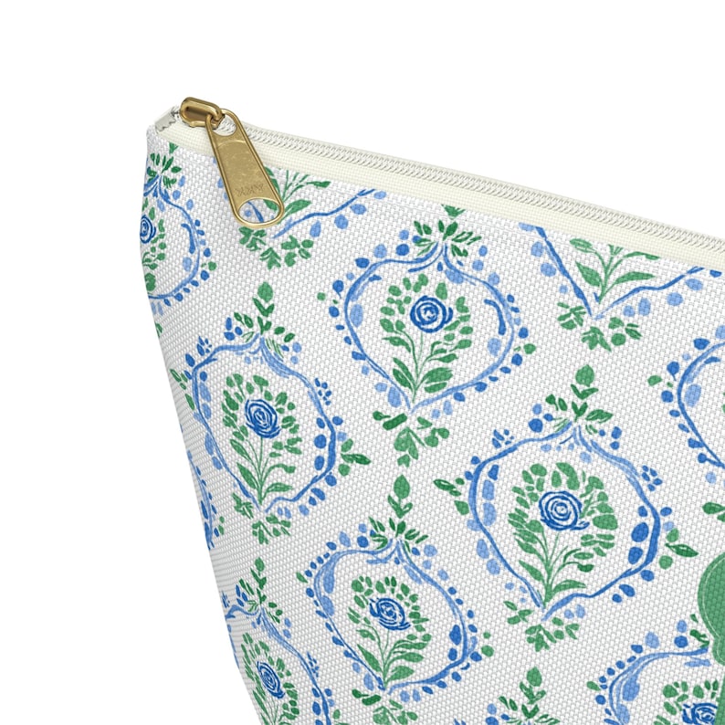 May include: A white cosmetic bag with a repeating pattern of blue and green floral designs. The bag has a gold zipper and a white interior. The floral pattern includes stylized roses and leaves.