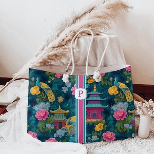 May include: A rectangular tote bag with rope handles featuring a dark teal background with a pattern of colorful peacocks, pink and yellow flowers, and pagodas. A vertical pink and teal stripe runs down the center with a white label and letter P.