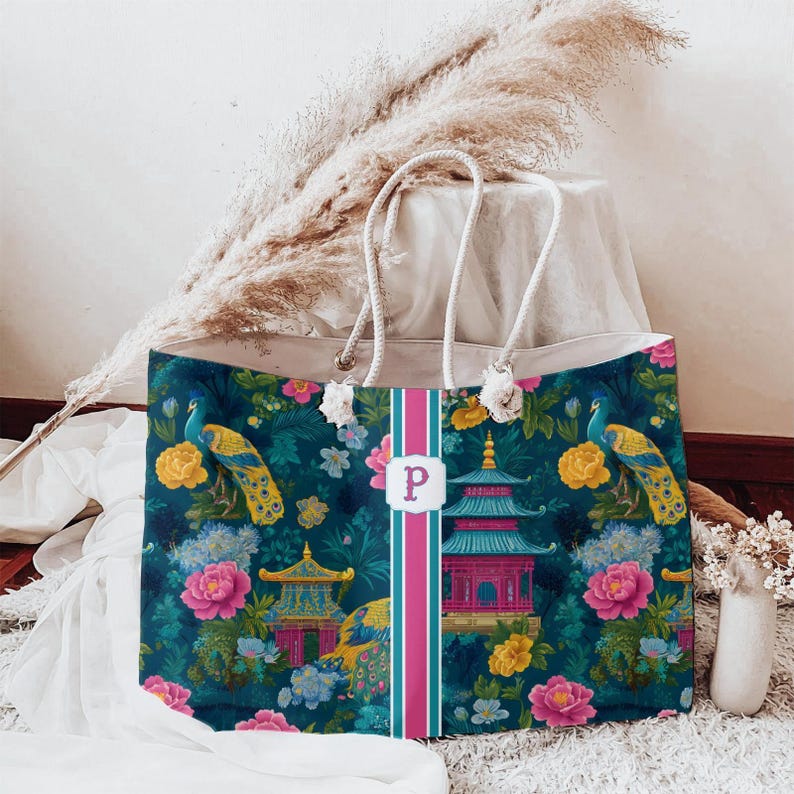 May include: A rectangular tote bag with rope handles featuring a dark teal background with a pattern of colorful peacocks, pink and yellow flowers, and pagodas. A vertical pink and teal stripe runs down the center with a white label and letter P.