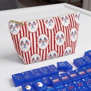 May include: A patriotic cosmetic bag with a red and white striped pattern and repeating flag designs. The bag has a gold zipper and is next to a set of blue Mahjong tiles with various symbols and numbers.