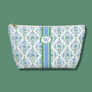 May include: A white cosmetic bag with a blue and green floral pattern. A green and blue stripe runs down the center, with a white oval containing the letter "W". The bag has a gold zipper.