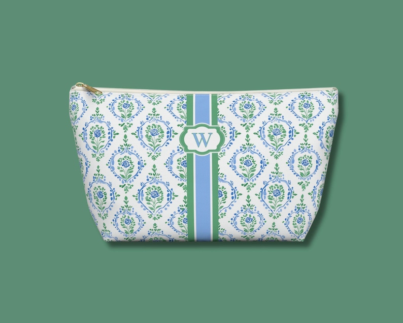 May include: A white cosmetic bag with a blue and green floral pattern. A green and blue stripe runs down the center, with a white oval containing the letter "W". The bag has a gold zipper.
