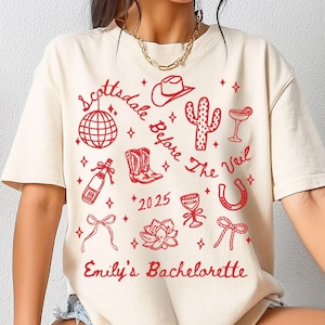 May include: A white t-shirt with a red graphic design featuring a cactus, cowboy hat, horseshoe, champagne bottle, and the text "Scottsdale Before The Veil 2025 Emily's Bachelorette".