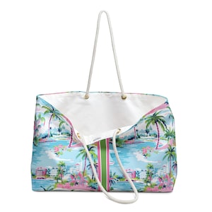 May include: A large tote bag featuring a light blue background with a tropical print of palm trees, pink flowers, and white buildings. The bag has a white interior, a vertical green and pink center stripe, and two white rope handles with gold grommets.