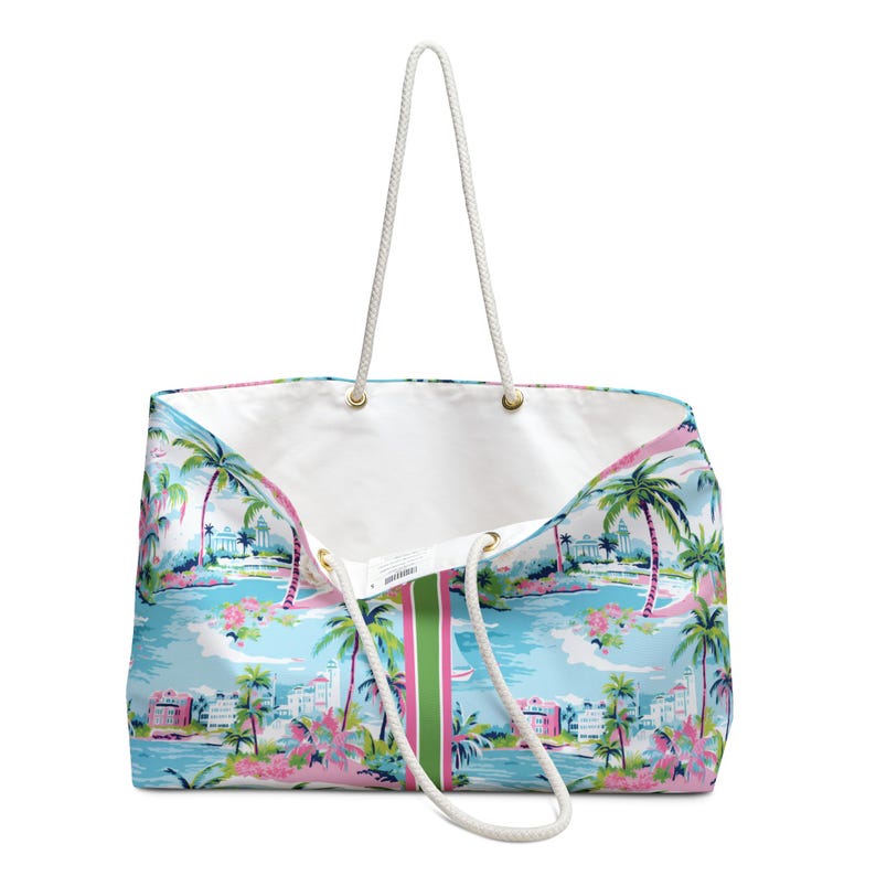 May include: A large tote bag featuring a light blue background with a tropical print of palm trees, pink flowers, and white buildings. The bag has a white interior, a vertical green and pink center stripe, and two white rope handles with gold grommets.