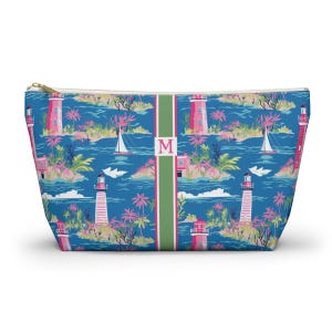 May include: A zippered pouch featuring a blue background with a repeating pattern of pink lighthouses, sailboats, and palm trees. A vertical green stripe runs down the center, displaying the letter M in a pink serif font on a white background.