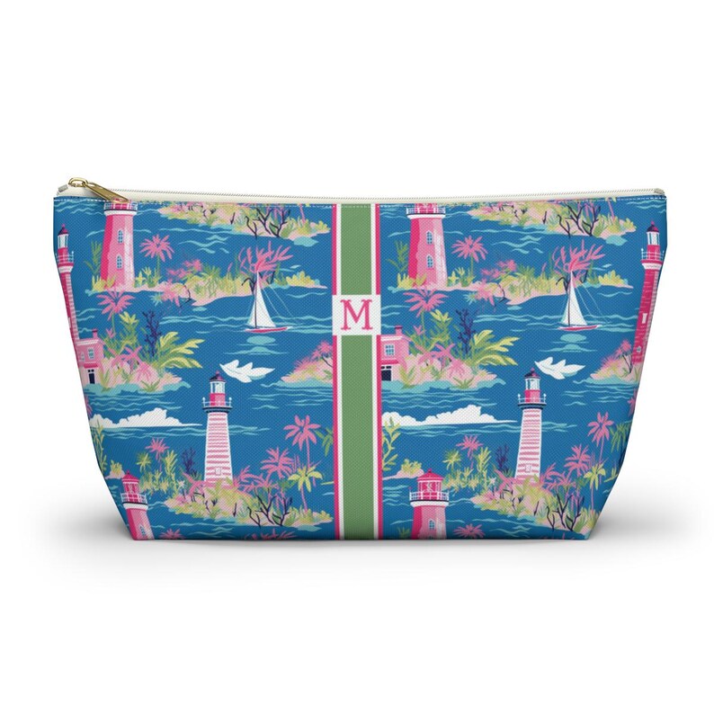 May include: A zippered pouch featuring a blue background with a repeating pattern of pink lighthouses, sailboats, and palm trees. A vertical green stripe runs down the center, displaying the letter M in a pink serif font on a white background.