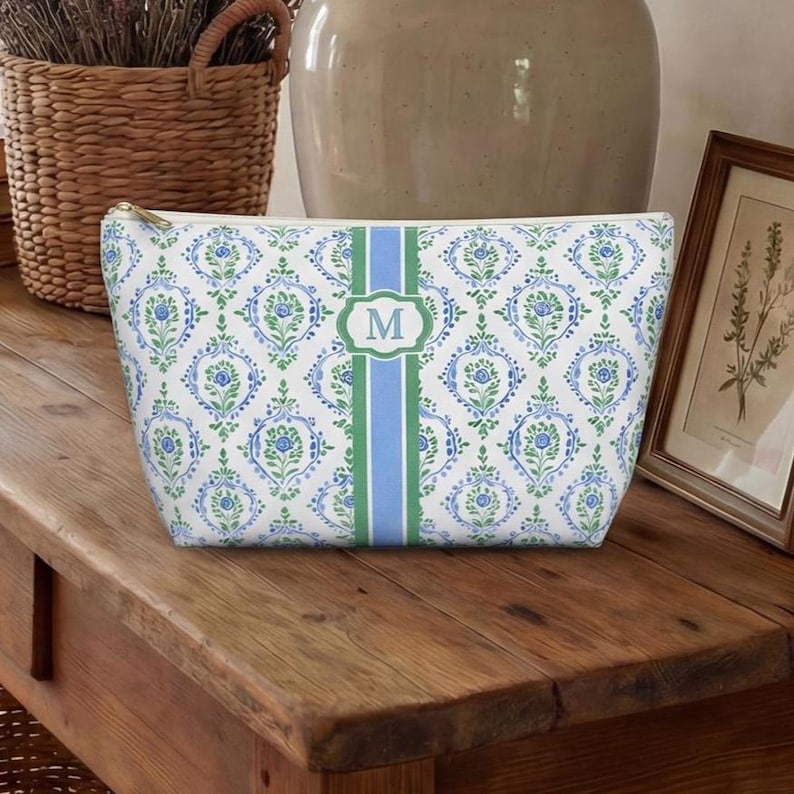May include: A white cosmetic bag with a blue and green floral pattern. The bag has a vertical stripe with the letter "M" in a green frame. The bag has a gold zipper and is sitting on a wooden surface.