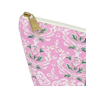 May include: A pink zippered pouch featuring a white damask pattern with floral motifs and green leaves. The bag has a gold-toned metal zipper pull attached to a white zipper track, showcasing a textured fabric surface with a repeating design.