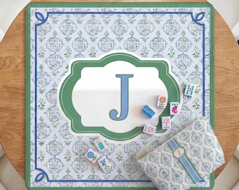 May include: A square mahjong mat featuring a light blue and green floral pattern with a central white scalloped emblem displaying the letter J in blue. Several small mahjong tiles are scattered on the mat alongside a matching zippered pouch.