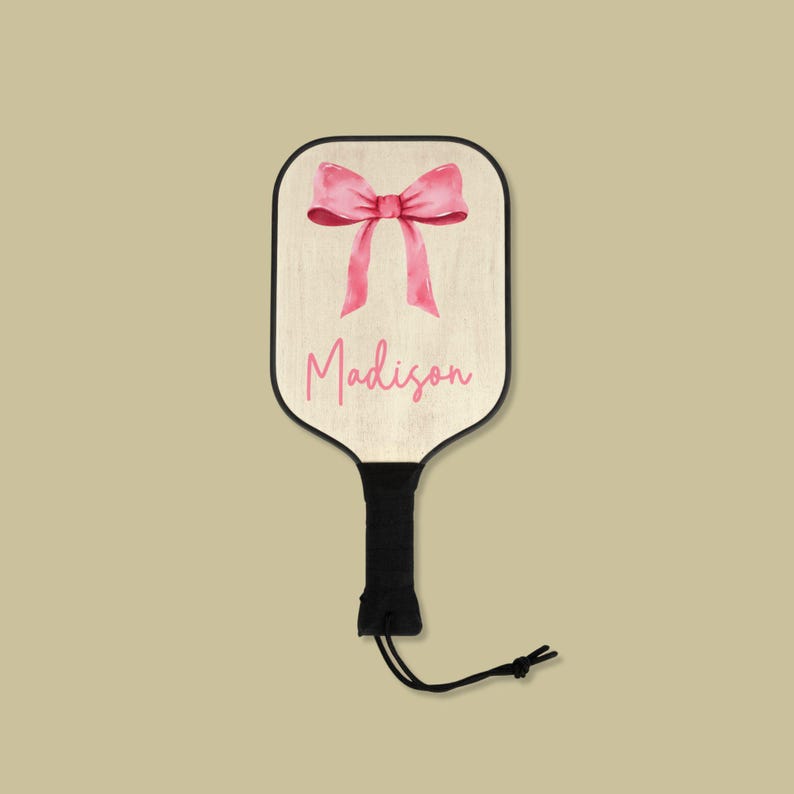 May include: A personalized wooden paddle with a pink bow and the name "Madison" printed on it. The paddle has a black handle with a string attached.