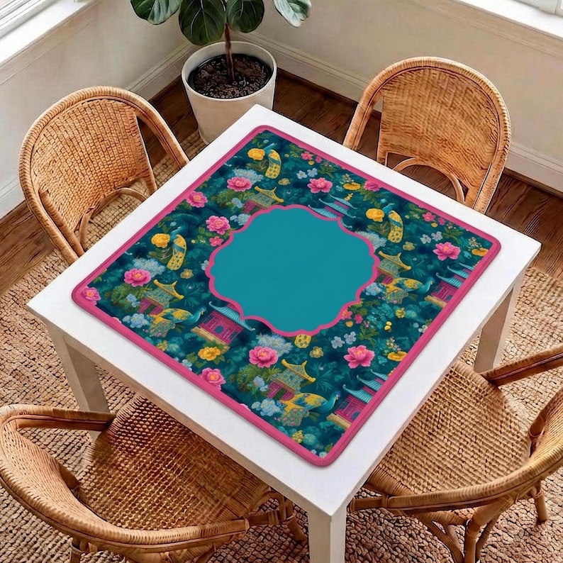 Op de afbeelding: Een vierkante tafelmat met een donkergroenblauwe achtergrond met een patroon van roze bloemen, gele vogels en kleine bouwwerken. De mat heeft een effen roze rand en een groot, centraal, blauwgroen kader met een geschulpte rand.