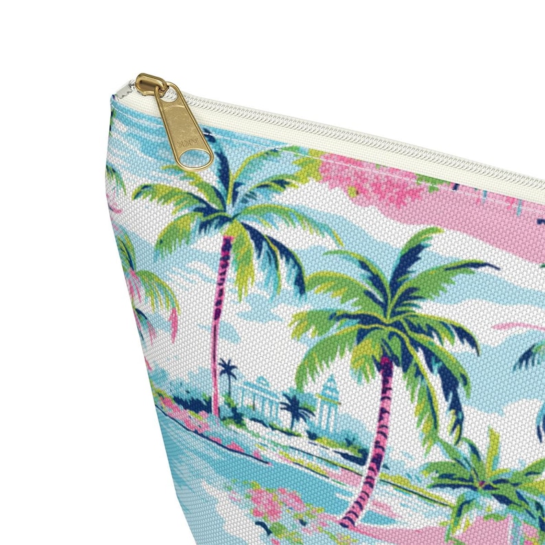 May include: A zippered pouch featuring a tropical print with green palm trees, light blue sky, and pink accents. The fabric has a visible woven texture, and the zipper pull is made of gold-toned metal, providing a contrast to the colorful pattern.