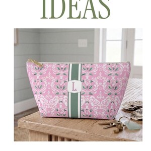 May include: A pink zippered pouch and a matching tote bag featuring a white damask floral pattern with a central green vertical stripe. Each bag displays a white rectangular monogram patch with a letter in the center and gold hardware.