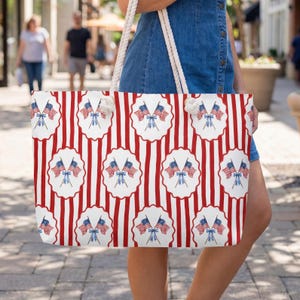 May include: A large tote bag with a patriotic design. The bag features a pattern of red and white stripes with repeating circular medallions. Each medallion displays two American flags and a blue bow. The bag has white rope handles.