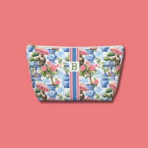 May include: A zippered cosmetic bag with a repeating pattern of monkeys, pink umbrellas, and blue and white vases. The bag has a vertical stripe with the letter "B" in the center. The background is a light pink.
