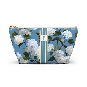 May include: A blue cosmetic bag with a floral pattern of white and light blue hydrangeas. The bag has a gold zipper and a vertical stripe with the letter "P".