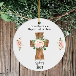May include: White ceramic Christmas ornament with a brown cross adorned with pink and white flowers and greenery. The ornament has the text "Saved by Grace Baptized In His Name", the name "Sydney", and the year "2023".