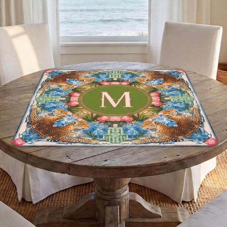 Puede incluir: Una alfombra decorativa cuadrada con un borde de estampado de leopardo y detalles florales. El centro presenta un &oacute;valo verde con la letra "M" en una fuente serif. La alfombra est&aacute; colocada sobre una mesa redonda de madera.