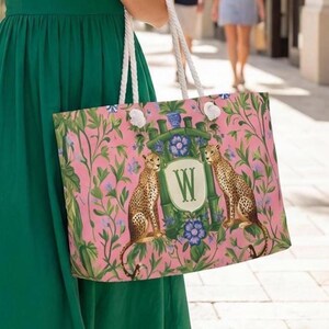 以下が含まれることがあります： 白いロープハンドルが付いたトートバッグで、ピンクの背景に緑の葉、青い花、そして中央のWの文字が入った紋章を挟むように2頭のチーターが描かれています。デザインには、中央のモノグラムエンブレムを囲む竹スタイルのフレームが含まれています。