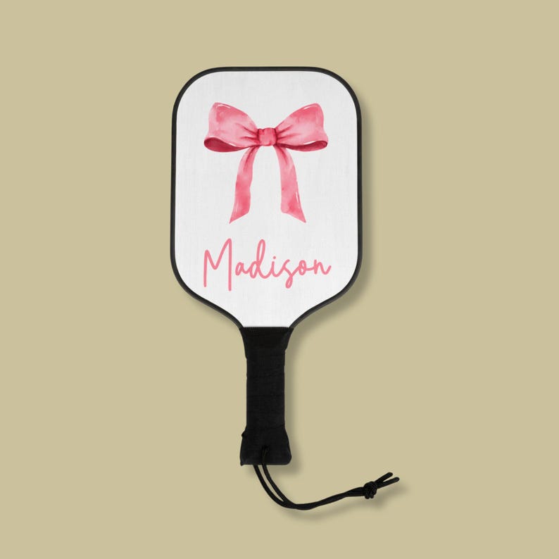 May include: A white paddle with a black handle and a pink bow with the name Madison written in pink cursive.