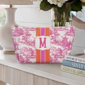 May include: A pink and white toiletry bag with a pink and orange stripe and the letter "M". The bag features a zipper closure and a landscape design. The bag is sitting on a white surface.