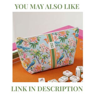 May include: A light blue zippered pouch featuring a floral and bird pattern with pink, orange, and blue colors. A vertical green and orange stripe runs down the center with the letter B printed in white. The pouch has a gold-toned metal zipper.