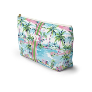May include: A rectangular zippered pouch featuring a vibrant tropical print with palm trees, coastal buildings, and pink floral accents. A vertical green and pink striped band runs down the center, displaying a white square with the letter S.
