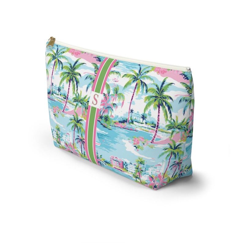 May include: A rectangular zippered pouch featuring a vibrant tropical print with palm trees, coastal buildings, and pink floral accents. A vertical green and pink striped band runs down the center, displaying a white square with the letter S.