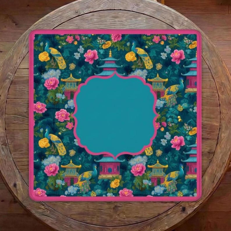 Op de afbeelding: Een vierkante placemat met een donkergroenblauwe achtergrond en een herhalend patroon van gele pauwen, roze pioenrozen en blauwe pagodes. De placemat heeft een effen middenstuk met een geschulpte roze rand en een bijpassende roze buitenrand.